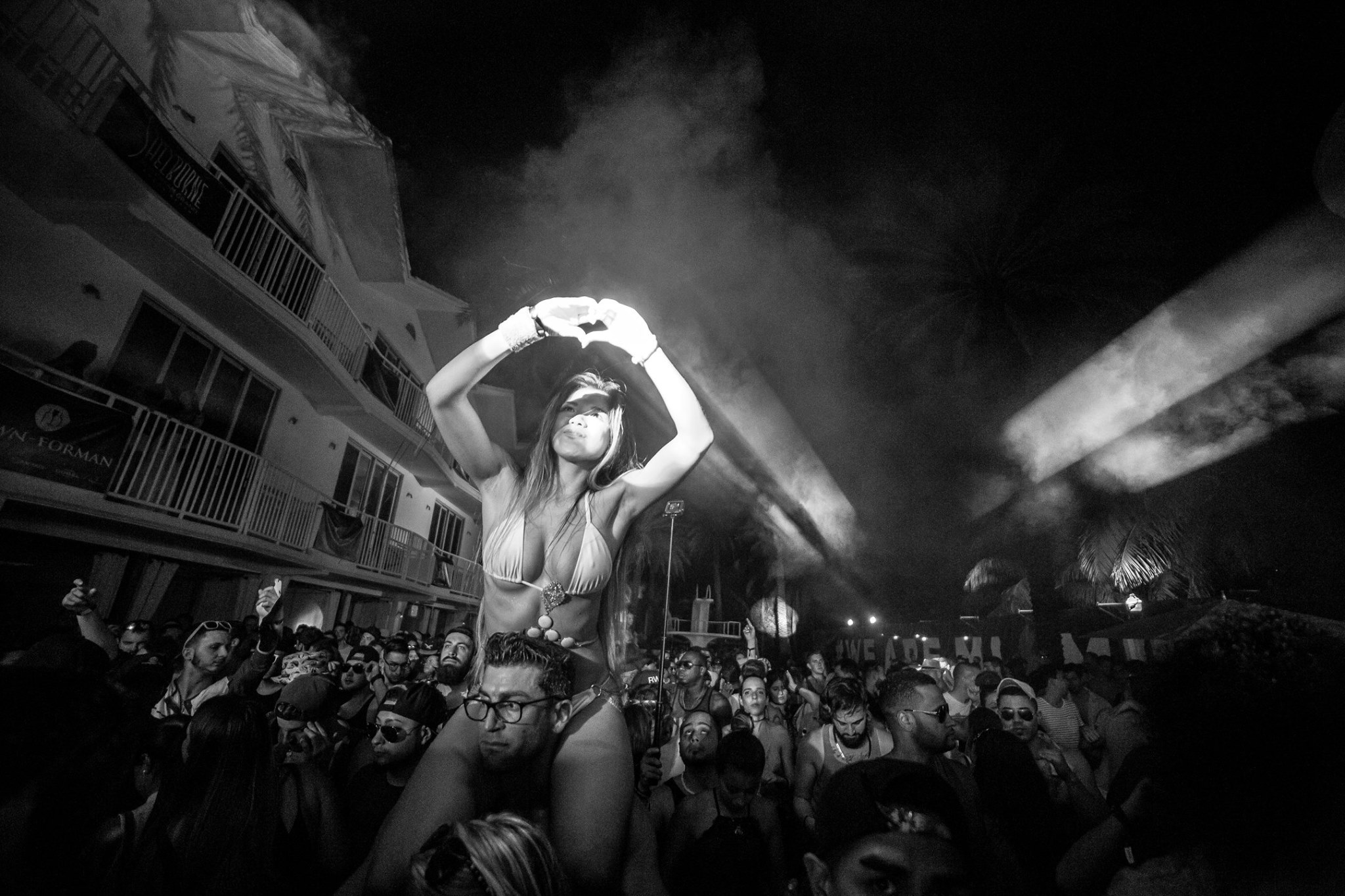 Black and white night shot of a guest on shoulders making a heart with her hands above a packed crowd