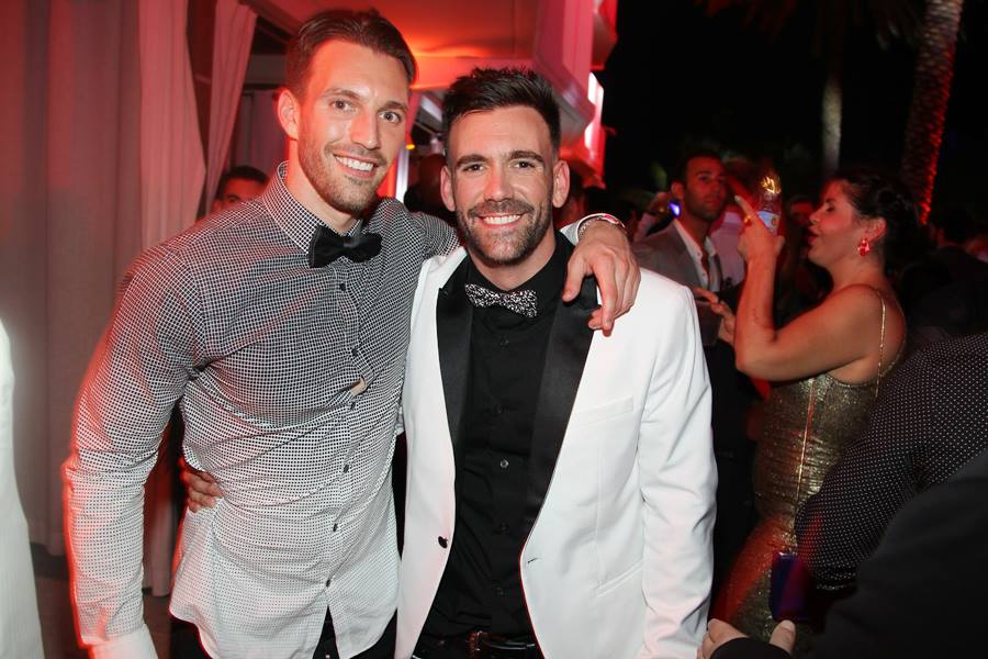 Two guests in black-tie formalwear, one in a white tuxedo jacket, posing on the dance floor under red light