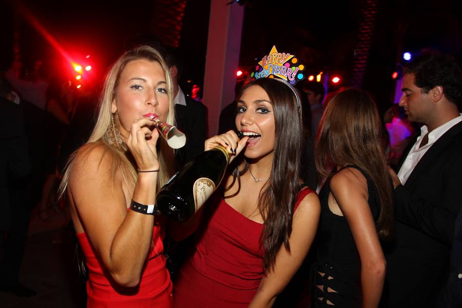 Two guests with party horns and champagne under pink light, one in a red dress and tiara