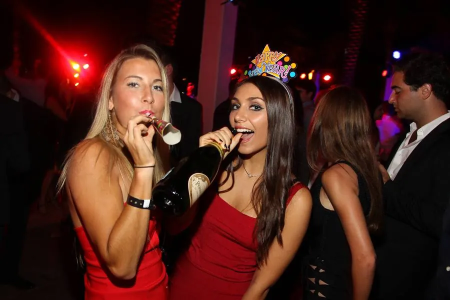 Two guests with party horns and champagne under pink light, one in a red dress and tiara
